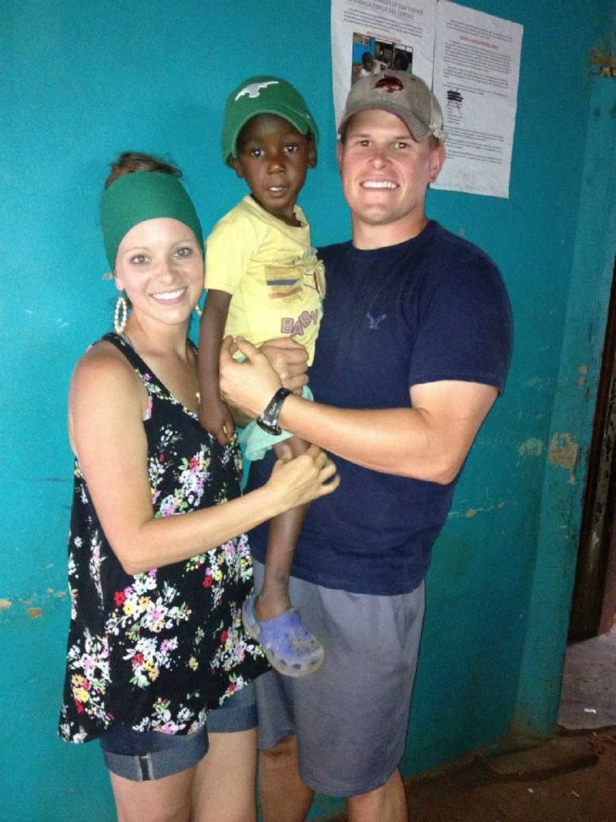 Husband and wife stand smiling while husband holds Ugandan orphan they are attached to