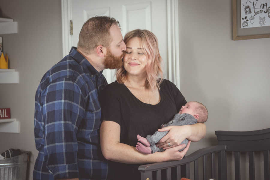 Husband kisses wife on cheek as she holds adopted newborn beside his crib in nursery
