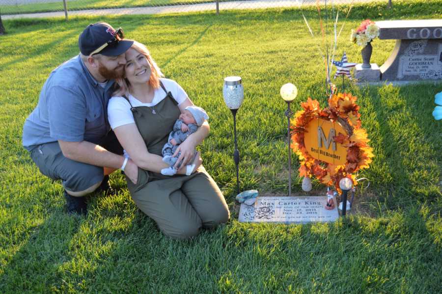 Mother kneels holding adopted newborn beside grave of their first born with husband squatting beside her