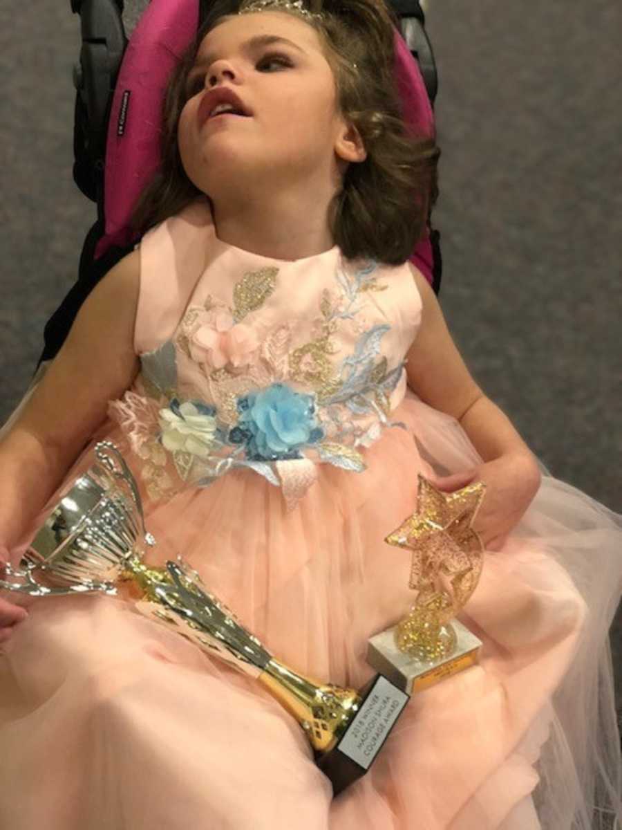 Little girl with shaken child syndrome sitting in stroller dressed for pageant with trophies on her lap