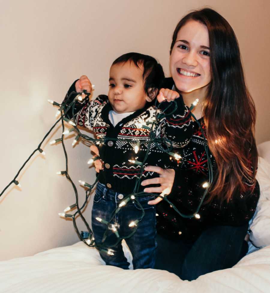 Single mother kneels behind baby son who is playing with strand of lights