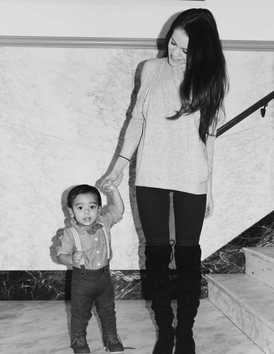 Mother stands holding hands with son who is in bow tie and suspenders