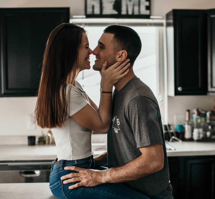 Girlfriend sits on kitchen counter smiling while holding boyfriend's face who stands in front of her