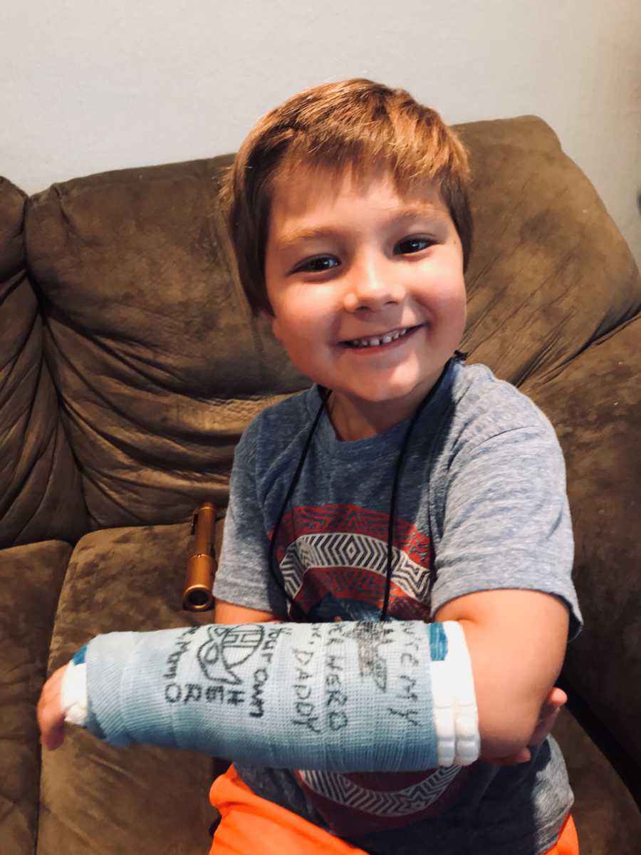 Little boy smiles as he sits on couch holding up blue cast on his arm with signatures all over it