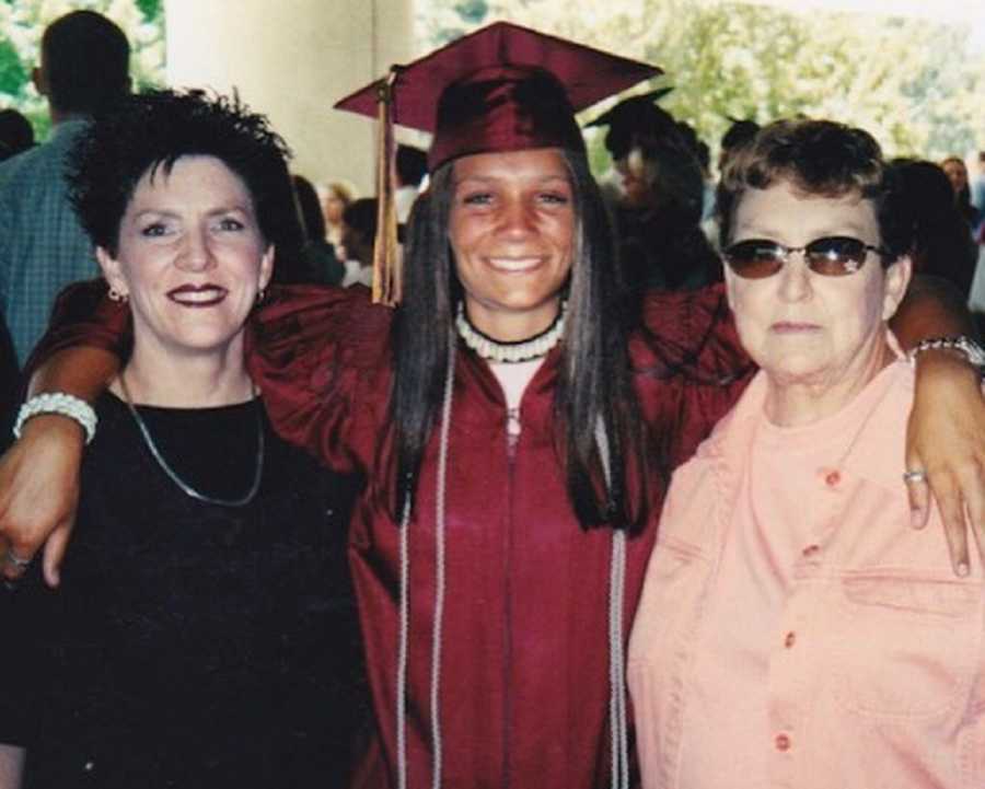 Teen in cap and gown stands smiling with arms around grandmother and mother who is drug addict