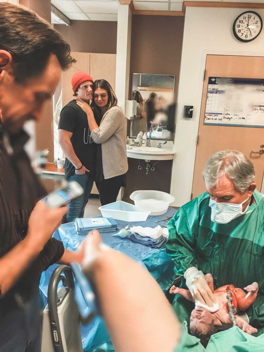 Husband and wife stand arm in arm as they they their newborn adopted baby in hospital room