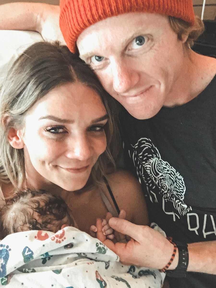 Newborn lays on adopted mother's chest while she smiles beside husband whose finger is being held by baby