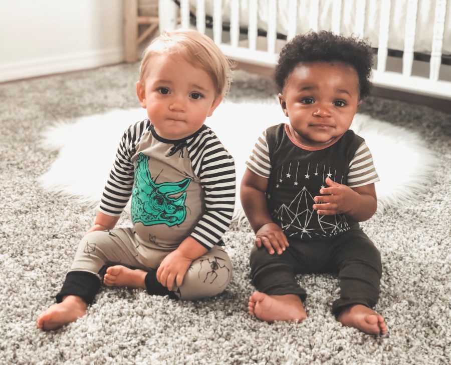 Newborn adopted brothers sit on carpet beside crib