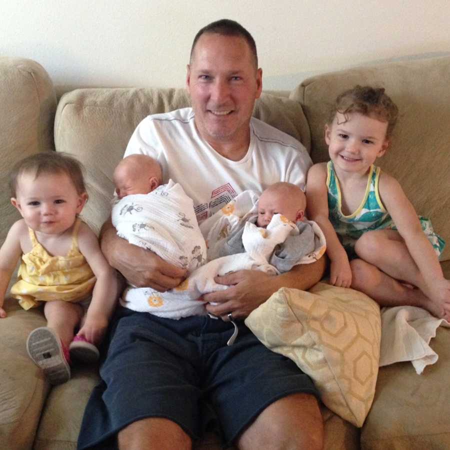 Man who has since passed sits on couch holding twin newborns with other two granddaughter sitting beside him