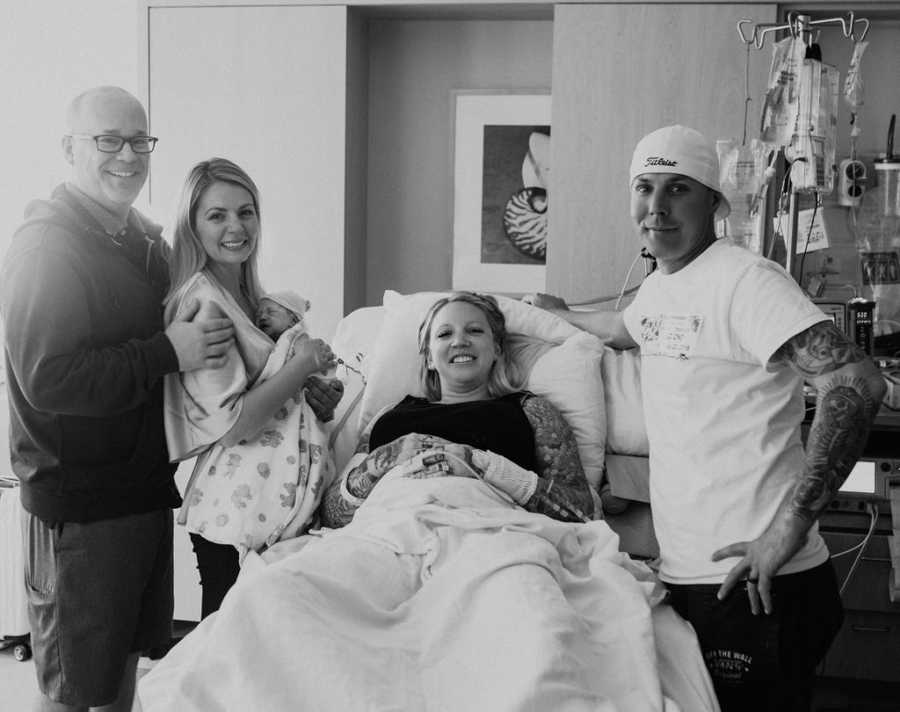 Husband and wife stand holding their newborn beside surrogate in hospital bed and her husband