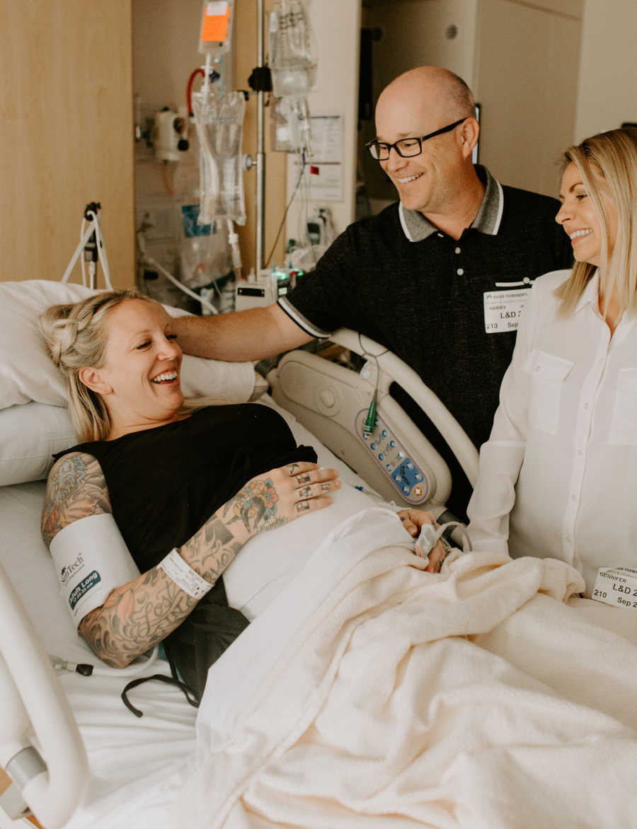 Parents of child stand beside pregnant surrogate laying in hospital bed