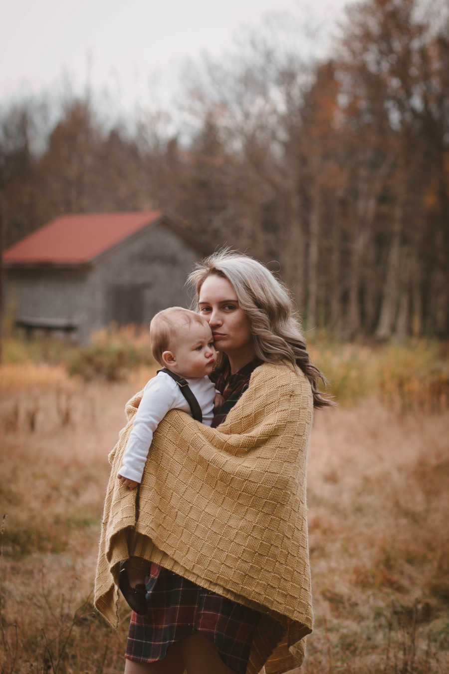 Mother stands in field holding baby son with blanket around her 
