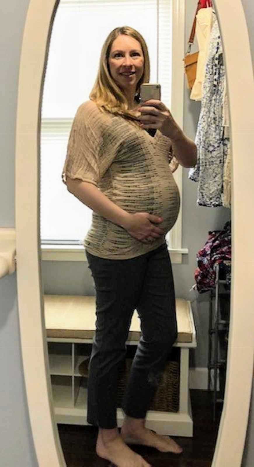Pregnant woman stands smiling while holding stomach in mirror selfie
