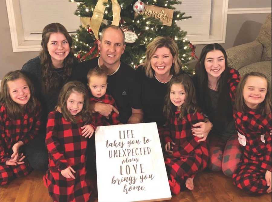 Family of nine sits on floor in front of Christmas tree with sign that says, "Life takes you to unexpected places..."