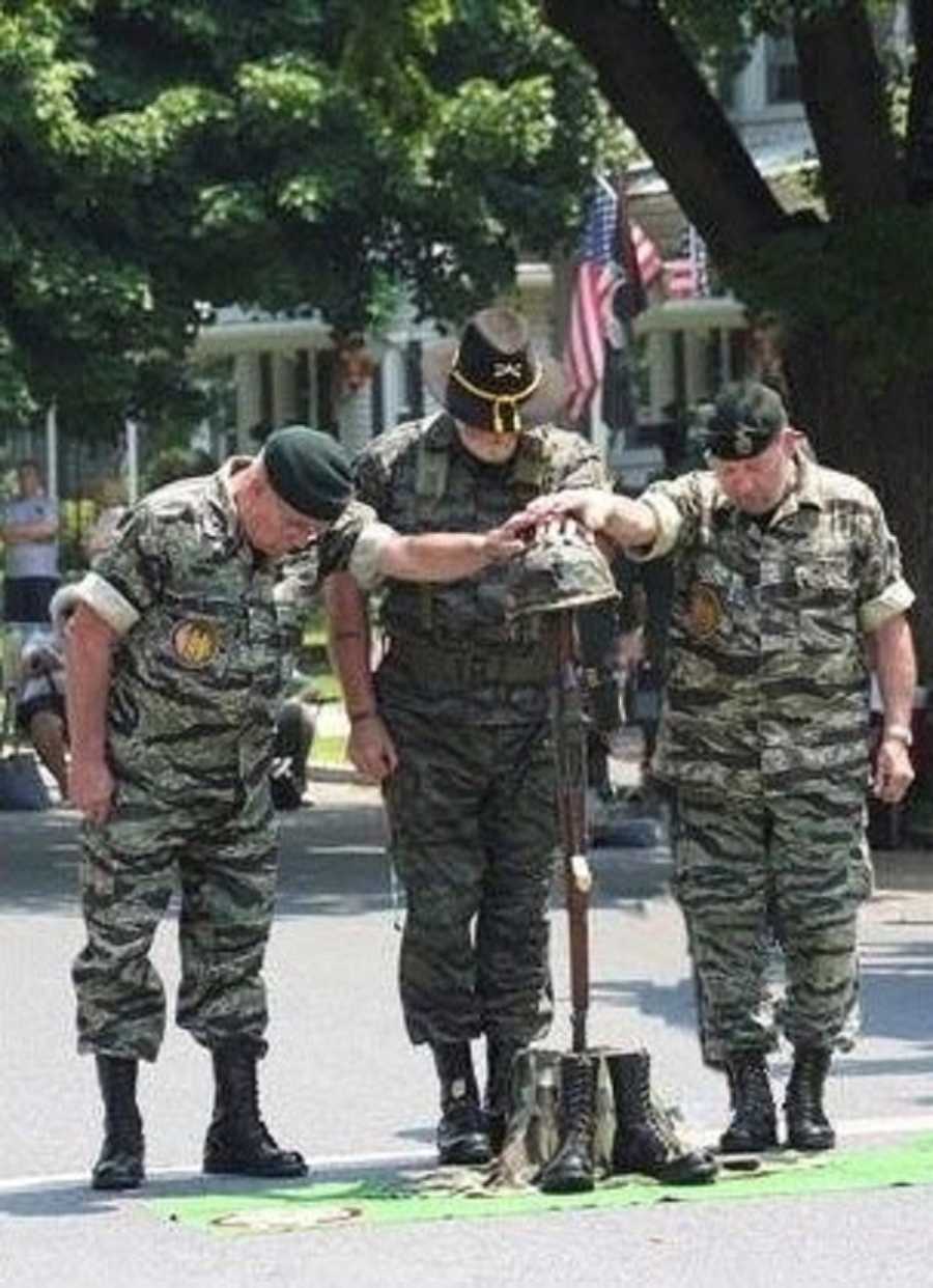Three vets stand in street conducting Field Cross with hands on fallen soldiers gun