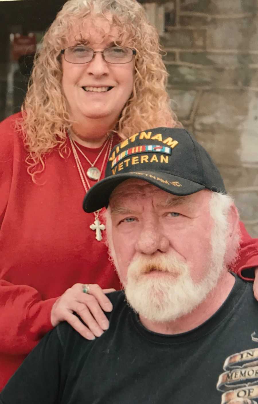 Vietnam veteran sits with his wife standing behind him with hands on his shoulders