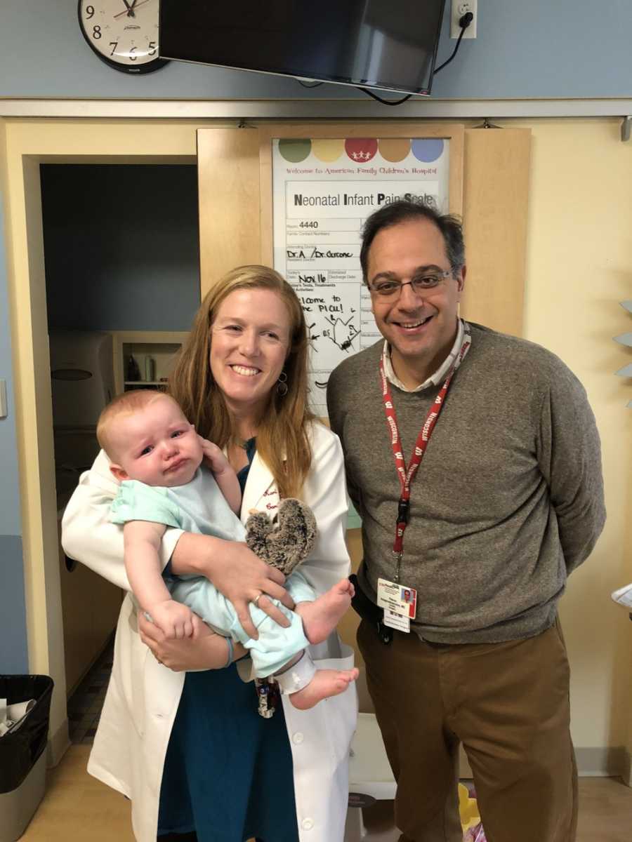 Two doctors stand smiling while one holds baby who need heart surgery