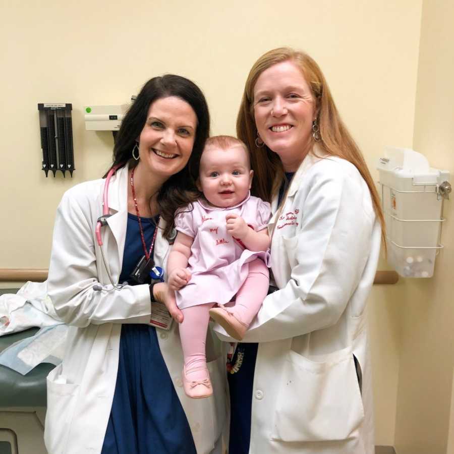 Baby girl with CHD smiles as she is held by two female doctors in doctors office