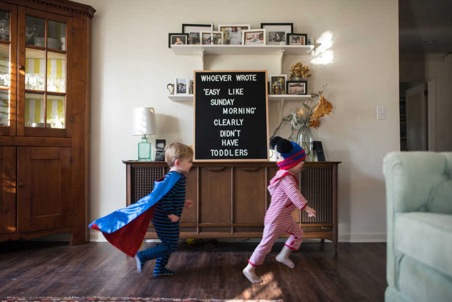 Young brother and sister chasing each other in house with sign in background that explains toddlers aren't easy