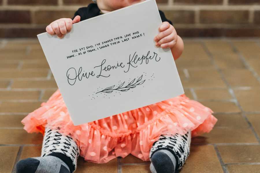 Little girl sitting on ground outside of adoption court holding sign that announces her adoption