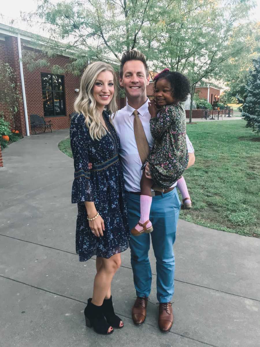 Husband and wife stand outside holding adopted toddler 
