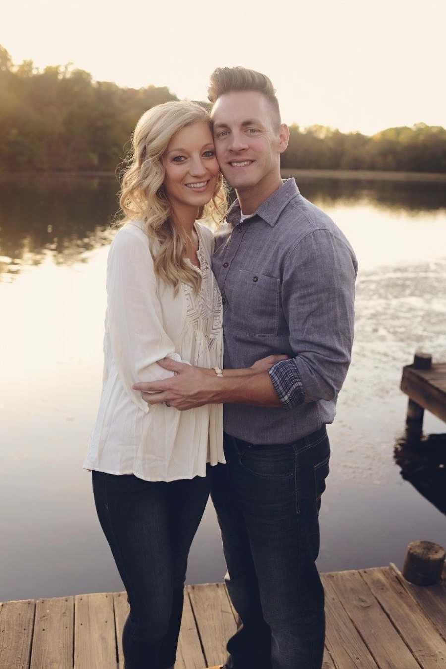 Husband and wife who struggle wit get pregnant stand arm in arm on dock beside body of water