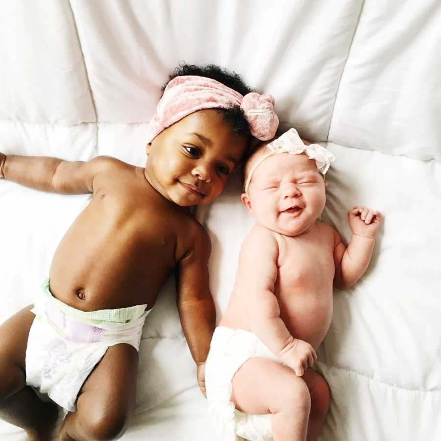 Woman's adopted baby and biological baby lie on their backs on white down comforter