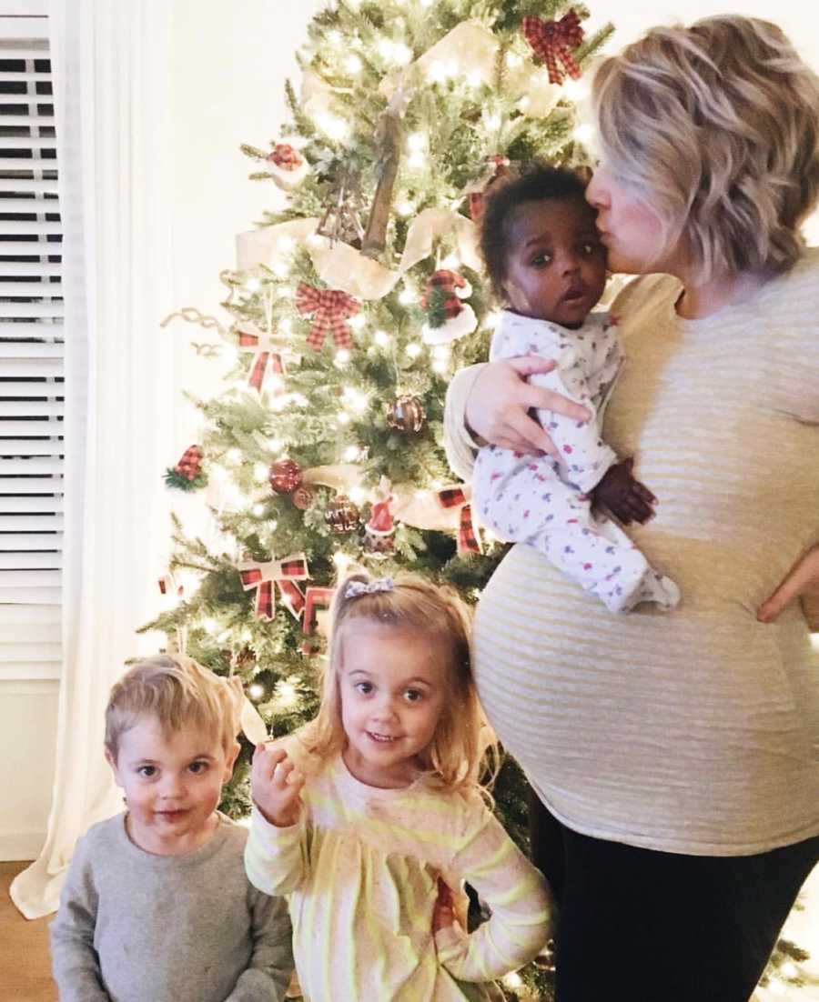 Pregnant mother holds adopted baby in her arms while biological son and daughter stand beside her in front of Christmas tree