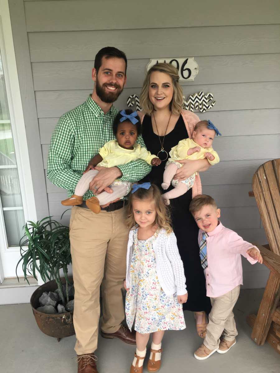 Husband and wife stand by their front door with their adopted baby, biological baby and son and daughter