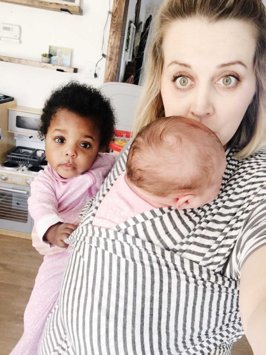 Woman stands while biological baby is swaddled to her chest and she holds adopted baby