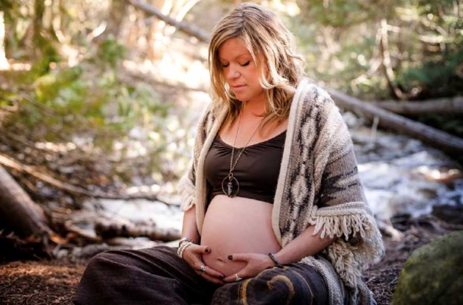 Pregnant woman sits on ground in woods holding stomach