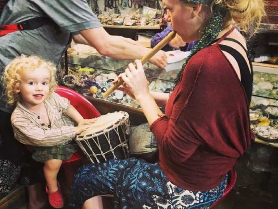 Mother sits playing flute while young son sits beside her playing the drums