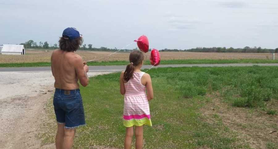 Shirtless father stands beside daughter outside as they hold onto red heart balloons
