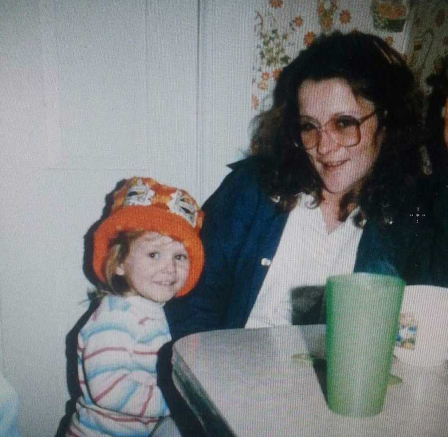 Little girl with orange hat on smiles while standing beside mother who sits at table