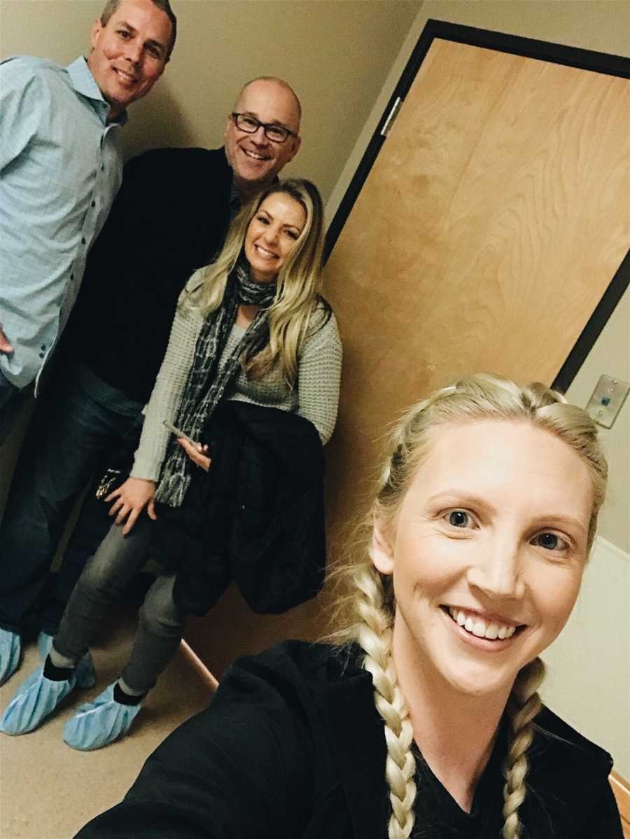 Surrogate smiles in selfie in doctor's office with husband and parents of baby she is carrying