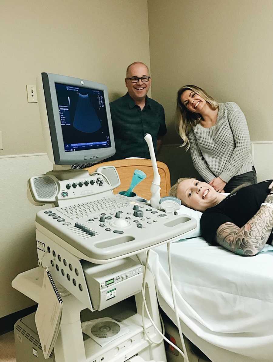 Surrogate laying down at ultrasound appointment with parents of baby standing beside her