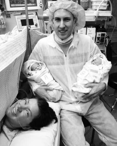 Woman smiles after undergoing c-section beside husband who sits in chair holding newborn twins