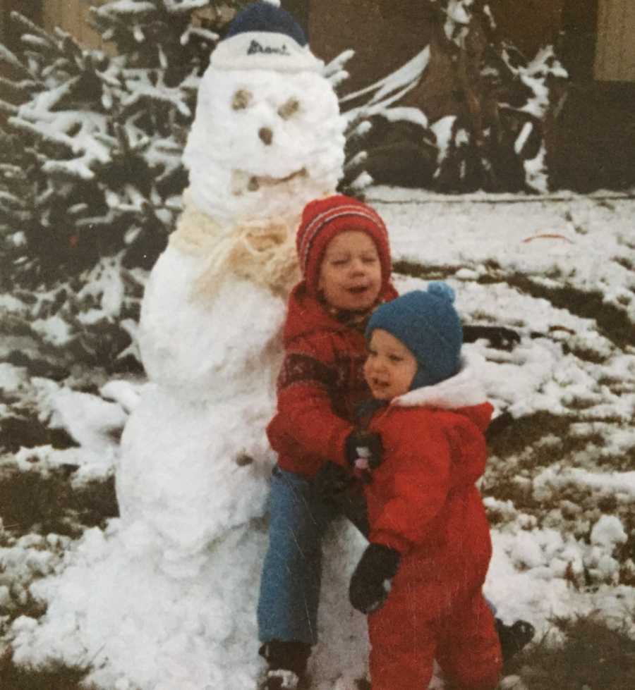 Young siblings stand outside in snow gear beside snow man