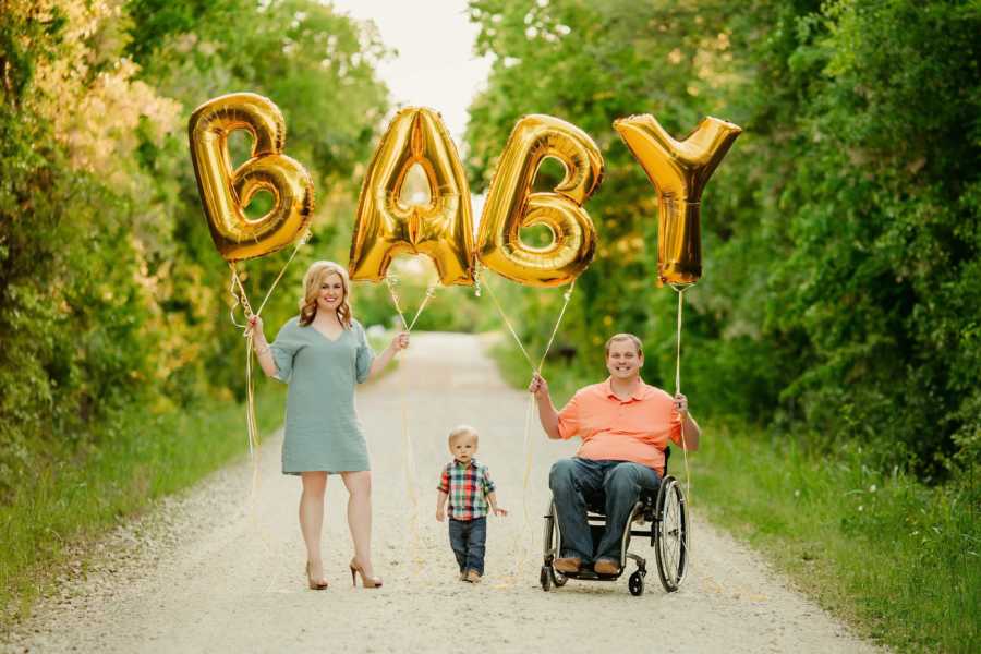 Pregnant woman and husband in wheelchair hold up balloons that spell out "baby" with their son standing between them