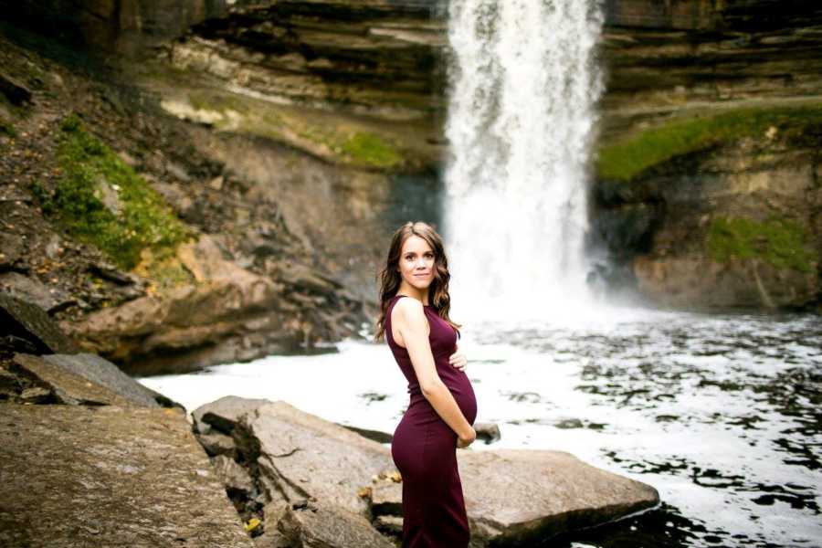 Pregnant woman standing in maroon dress by waterfall