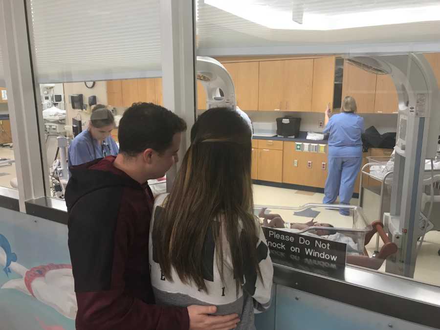 Husband and wife standing beside glass wall in hospital watching their adopted newborn