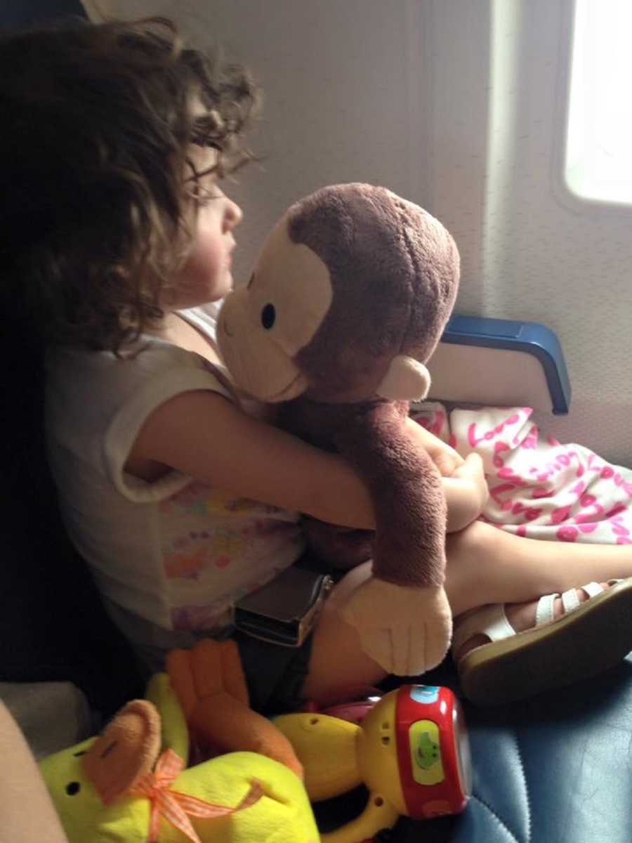 Little girl sitting in airplane looking out window while holding Curious George stuffed animal