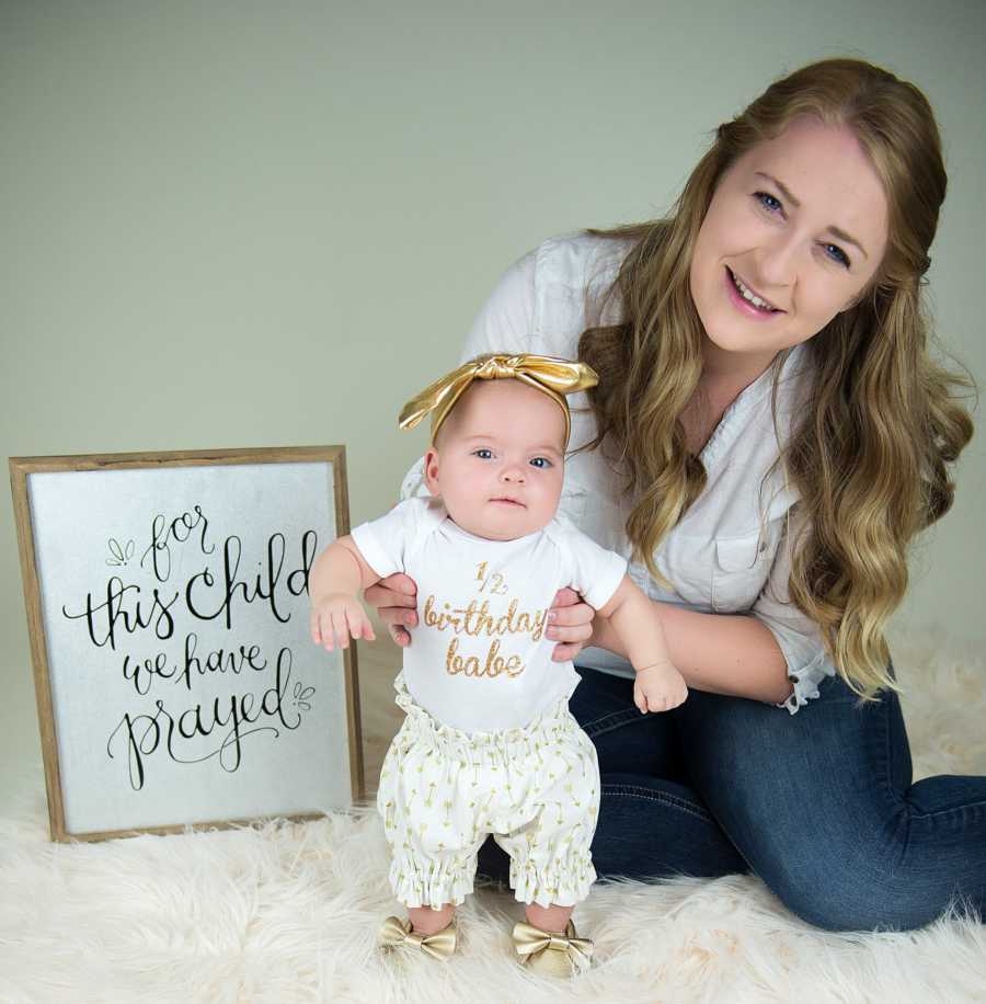 Adopted mother sits on white rug holding up baby who stands beside sign that says, "For this child we have prayed"