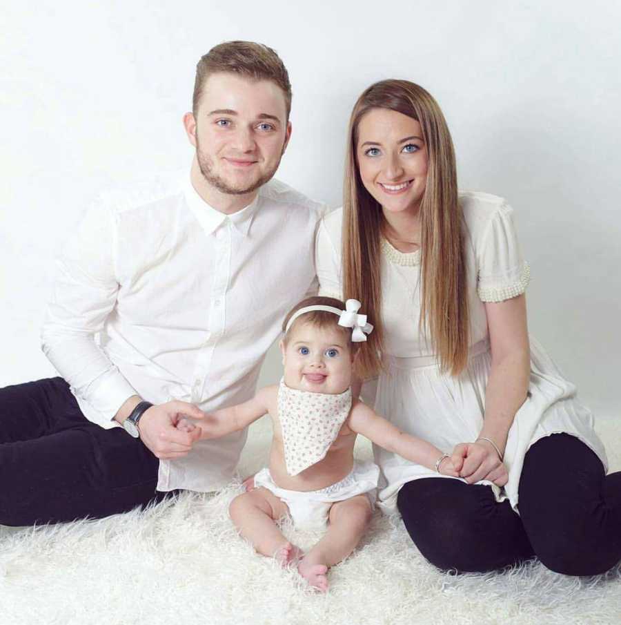 Husband and wife sit on white carpet beside baby daughter with cystic hygroma for photoshoot
