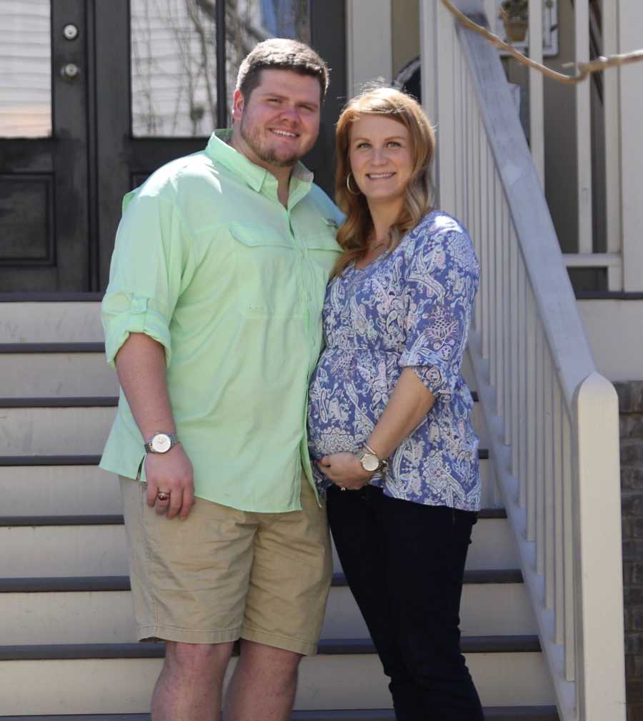 Pregnant wife smiles on outdoor steps of home beside husband 