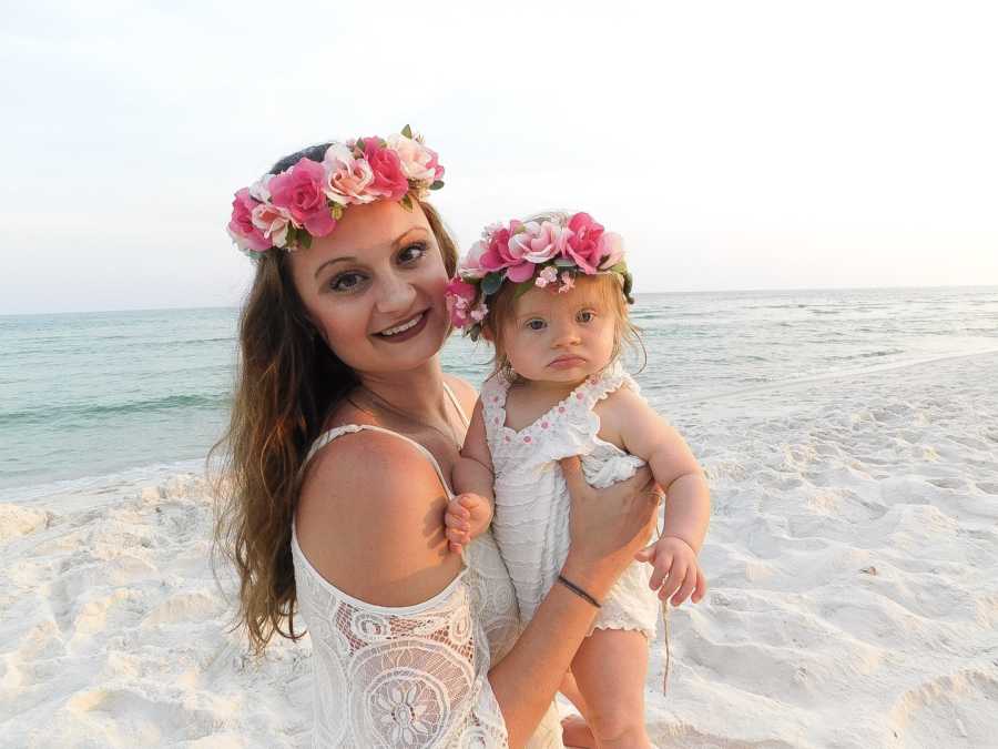 Mother stands on beach holding baby girl with down syndrome as they wear matching pink flower crowns