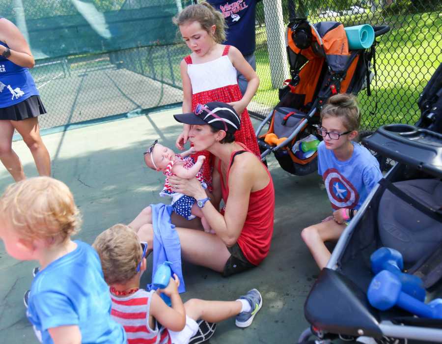Mother sits on tennis court holding baby with four other children surrounding her