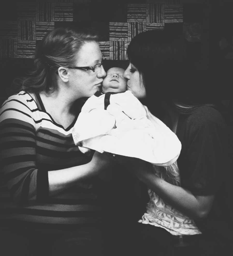 Birth mother and adoptive mother sit kissing the cheeks of newborn 