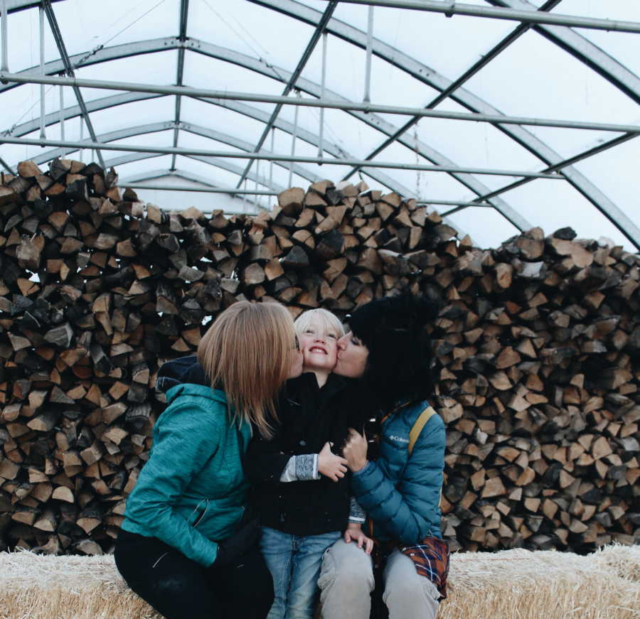 Adoptive mother and birth mother sit on hay bale kissing cheeks of their son