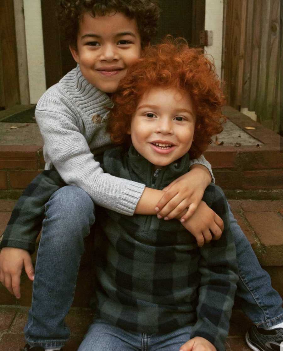 African American boy with red hair sits on steps outside of home while older brother sits behind him with his arms around him